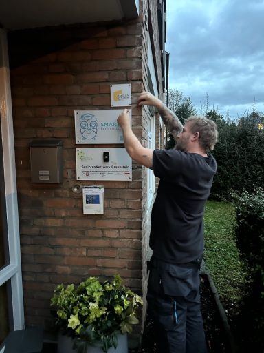 Mann bringt ein Schild an die Wand vor einem Gebäude an. Umgeben von Pflanzen und Gras.
