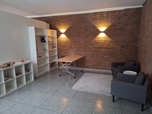 Heller Raum mit Ziegelwänden, einem Tisch, zwei grauen Sesseln und Regalen.